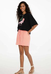 Žena s dlouhými kudrnatými vlasy, usmívající se, oblečená v černém oversized graphic tričku, oranžových a bílých pruhovaných šortkách a béžových sandálech se šněrováním.