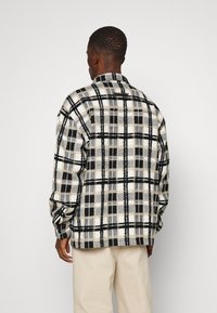 Camisa de flanela em padrão xadrez nas cores preto, branco e bege; possui colarinho largo, mangas longas e um corte descontraído, com punhos abotoados e yoke nas costas.