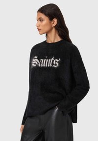 Musta pörröinen poolopaita, jonka etupuolella on näytt�ävä valkoinen "Saints"-teksti. Pyöreä kaula-aukko ja rento siluetti, yhdistetty mustiin nahkahousuihin.