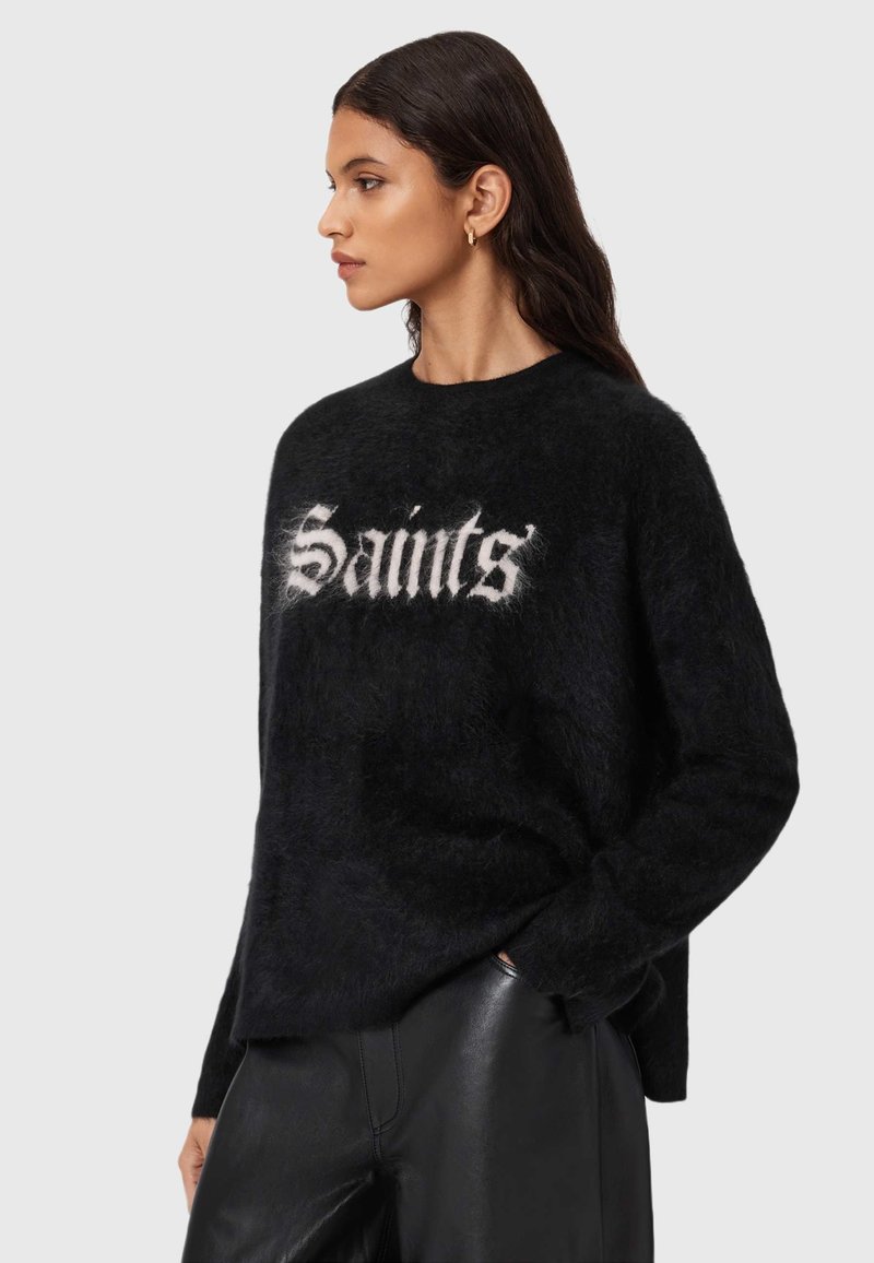 Musta pörröinen poolopaita, jonka etupuolella on näyttävä valkoinen "Saints"-teksti. Pyöreä kaula-aukko ja rento siluetti, yhdistetty mustiin nahkahousuihin.