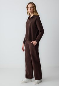 Pullover in pile marrone con colletto a mezza zip, abbinato a pantaloni larghi coordinati. Presenta tasche frontali e sneakers bianche.