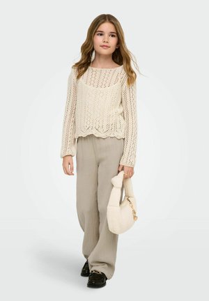 Młoda dziewczyna z długimi falowanymi włosami, ubrana w kremowy sweter ażurowy, beżowe szerokie spodnie, czarne mokasyny, trzymająca puszystą białą torebkę.