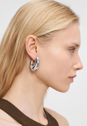 Primo piano di una donna che indossa un grande orecchino a cerchio curvo in argento lucido nell'orecchio sinistro, con capelli biondi lisci e una maglietta senza maniche marrone.