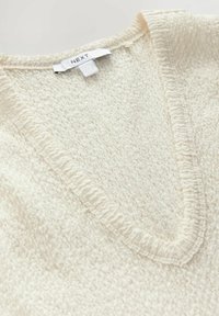 Cremfarbener Pullover mit V-Ausschnitt, strukturiertem Stoff und gerippten Details entlang des Ausschnitts und des Saums. Markenlabel ist sichtbar.