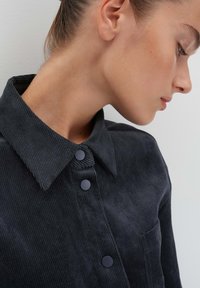 Camicia in corduroy nera con colletto strutturato, due bottoni neri e tasca sul petto con texture a coste.