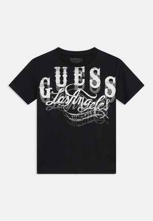 Čierne tričko s krátkym rukávom s veľkým bielym a sivým vrstveným nápisom „GUESS Los Angeles“ na prednej strane.