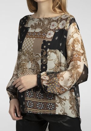 Kvinde iført bluse med lange ærmer med blandet blomster- og geometrisk patchworkmønster i beige, sort og brun nuancer.