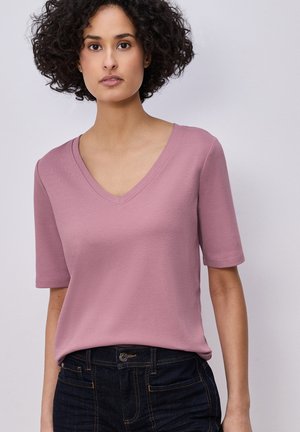 Frau mit lockigem dunklem Haar, die ein mauvefarbenes V-Ausschnitt-T-Shirt mit kurzen Ärmeln und dunkle Jeans aus Denim trägt, stehend vor einem schlichten hellen Hintergrund.