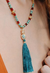 Collier en perles avec des accents turquoise, rouge et or ; il présente un pendentif en or avec une pierre turquoise et un pompon teal.