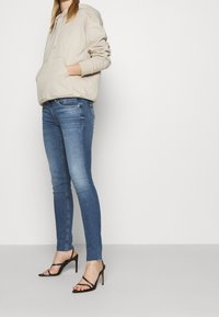 Sudadera de algodón beige con bolsillo tipo canguro, combinada con jeans ajustados de color azul oscuro y tacones altos negros con tiras. Conjunto simple y casual.