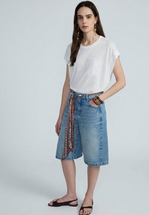 Donna con camicia bianca a maniche corte, pantaloncini di jeans fino al ginocchio con cintura a fantasia, numerosi braccialetti e sandali scuri aperti sulla punta, in piedi davanti a uno sfondo grigio.