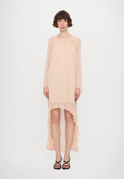 Pfirsichfarbnes, langärmeliges Kleid mit Rüschensaum am Hals und am Saum, im High-Low-Design, aus leichtem Stoff, getragen mit schwarzen Sandalen mit Riemchen.