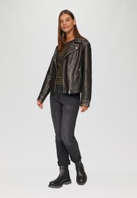 Zwarte leren motorjack met zilveren details, gestreept olijf- en zwart shirt, zwarte spijkerbroek en robuuste enkelboots. Model staat in profiel.