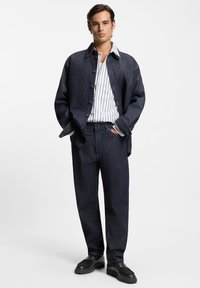 Giacca di jeans blu scuro, abbinata a una camicia bianca a righe blu navy e jeans a gamba larga blu scuro. Scarpe nere con suola spessa.