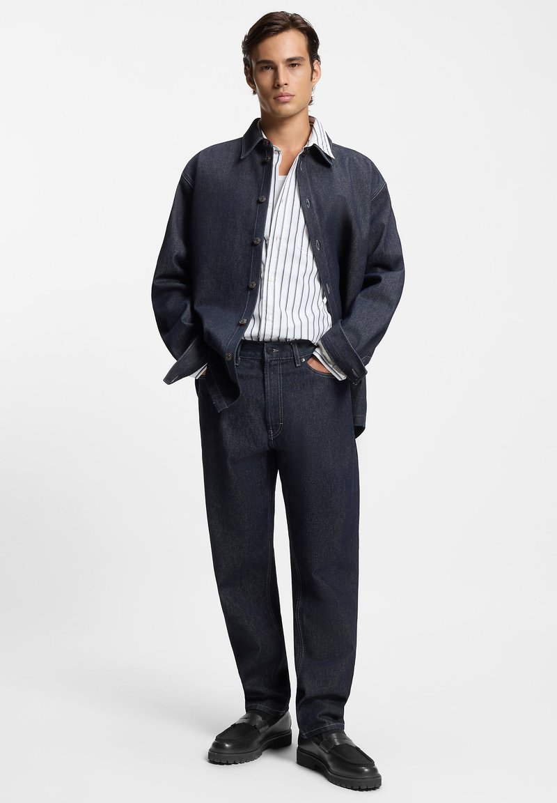 Giacca di jeans blu scuro, abbinata a una camicia bianca a righe blu navy e jeans a gamba larga blu scuro. Scarpe nere con suola spessa.