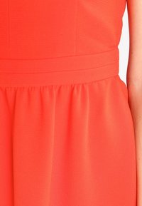 Korálové šaty s texturovanou látkou, s přiléhavým trupem, širokým páskem a rozšířenou sukní s nařasením.