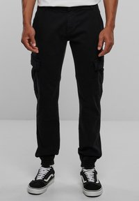 Pantalon cargo noir avec poignets élastiques aux chevilles, doté de poches latérales et d'une texture en coton lisse. Assorti à des baskets noires et blanches.