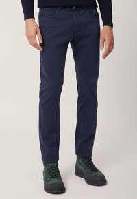 Pantaloni in cotone blu navy, slim-fit, con cinque tasche, dettagli di cucitura discreti, abbinati a stivali da trekking verde scuro.