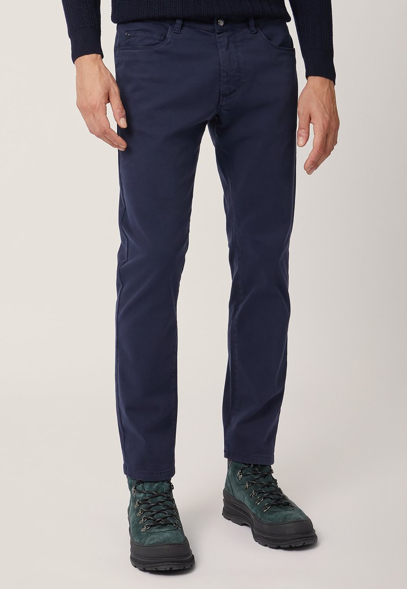 Pantaloni in cotone blu navy, slim-fit, con cinque tasche, dettagli di cucitura discreti, abbinati a stivali da trekking verde scuro.