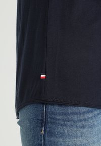 Dolní část těla v černém tričku s malou červenobílo-modrou nášivkou nad modrými džíny.