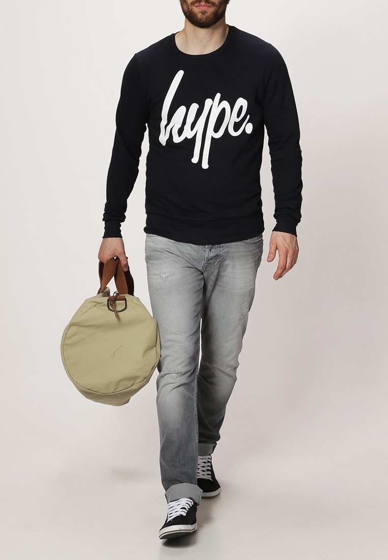 Homme marchant portant un sweat-shirt noir "hype.", jean gris, baskets noires, portant un sac rond beige avec des poignées marron.