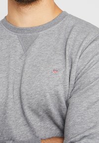 Šedá mikina s kulatým výstřihem, žebrovanými manžetami a malým vyšitým logem na hrudi. Vyrobena z měkké, strukturované tkaniny.