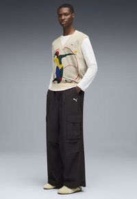 Chaleco de punto con un diseño abstracto colorido sobre una camiseta de manga larga blanca; combinado con pantalones cargo negros holgados y zapatos de color claro.