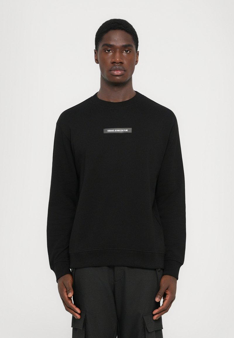 Svart sweatshirt med rund hals og ribbestrikkede mansjetter; har et sentralt hvitt merke med teksten "VERSACE JEANS COUTURE". Mykt bomullsmateriale.