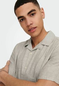 Camisa gris claro de manga corta con cuello abotonado, hecha de un tejido texturizado. Presenta botones en la parte delantera y sutiles detalles de costura.