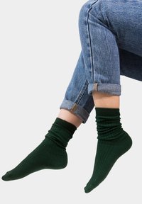 Ciemnozielone, prążkowane skarpetki z falbaniastym wykończeniem, noszone z podwiniętymi niebieskimi dżinsami. Materiał wydaje się miękki i elastyczny, o gładkiej fakturze.