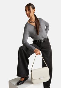 Grauer Pullover, schwarze weit geschnittene Jeans, schwarze Ballerinas, hält eine weiße strukturierte Handtasche mit silberner Hardware. Model posiert auf einem weißen Podest.