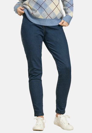 Person iført mørkeblå slim-fit jeans, hvide sneakers og en lyseblå argyle sweater med hænder, der justerer sweaterens kant.
