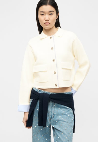 Jeune femme portant une veste crème boutonnée avec des boutons dorés, des poignets rayés bleus, un jean bleu orné de perles et un pull marine noué autour de la taille.