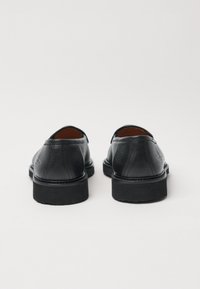 Černé kožené boty s texturovaným povrchem, pohled zezadu ukazuje zaoblené měkké okraje a nízkou gumovou podrážku. Minimalistický design, žádné viditelné kovové prvky.