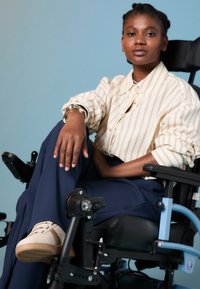 Elektrische rolstoel met een zwarte zitting en armleuningen, blauw frame en verstelbare voetsteunen. Onderwerp draagt een gestreept shirt, marineblauwe broek en beige schoenen.