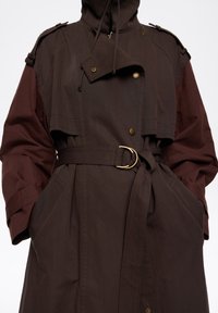 Płaszcz trencz z paskiem w kolorze ciemnobrązowym i bordowym, z wysokim kołnierzem, zatrzaskami i teksturowaną tkaniną. Pasek z klamrą typu D uzupełnia talię.