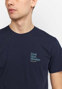Marineblauwe katoenen T-shirt met een print op de linkerborst: "Fresh Meat Mondays" in teal en gouden tekst. Ronde hals, korte mouwen, gladde oppervlakte.