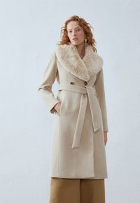 Cappotto in lana beige con colletto in pelliccia, chiusura doppiopetto, cintura in vita e maniche lunghe. Texture liscia, design minimalista.