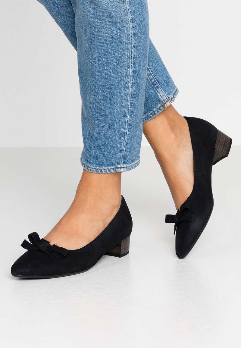 Pieds portant des chaussures en daim noir à petit talon avec des nœuds, associés à un jean en denim bleu raccourci sur un fond clair uni.
