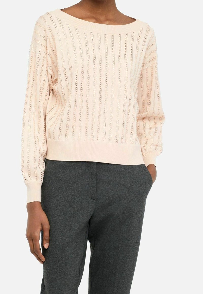 Femme portant un pull en maille côtelée rose clair avec des rayures verticales ajourées et un pantalon gris foncé, main dans la poche.