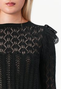 Primo piano di una donna che indossa un top in pizzo nero con motivi floreali e geometrici, con un dettaglio arricciato sulla spalla.