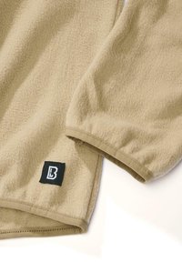 Bézs színű, puha tapintású polár pulóver, amelyen a szegélyen egy fekete címke található fehér logóval, finom mandzsetta részletekkel.