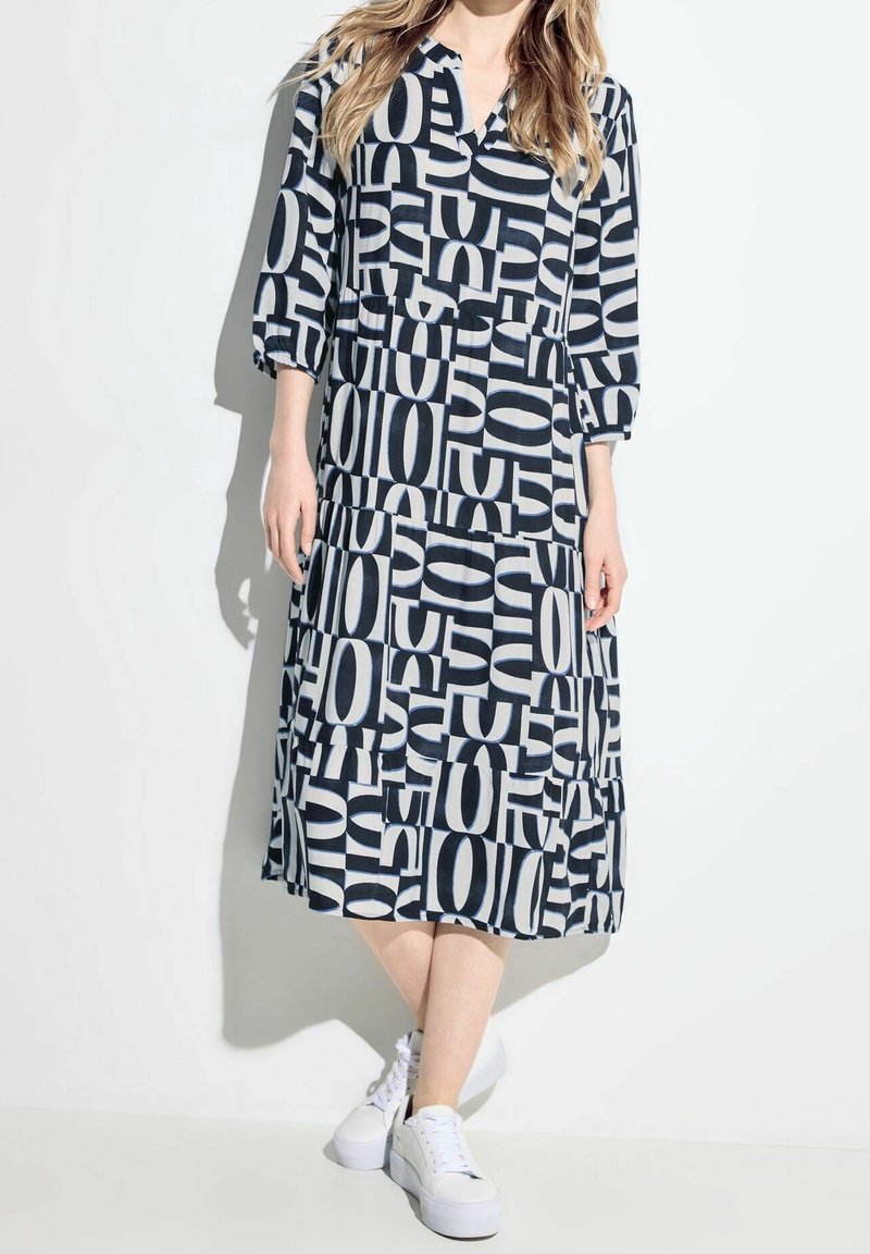 Femme portant une robe noire et blanche longueur genou avec des motifs abstraits de lettres, manches trois-quarts et baskets blanches à plateforme.