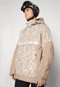 Mand iført beige og hvid camouflagemønster Burton-jakke med hætte og sort skihjelm med goggles, der kigger til siden.