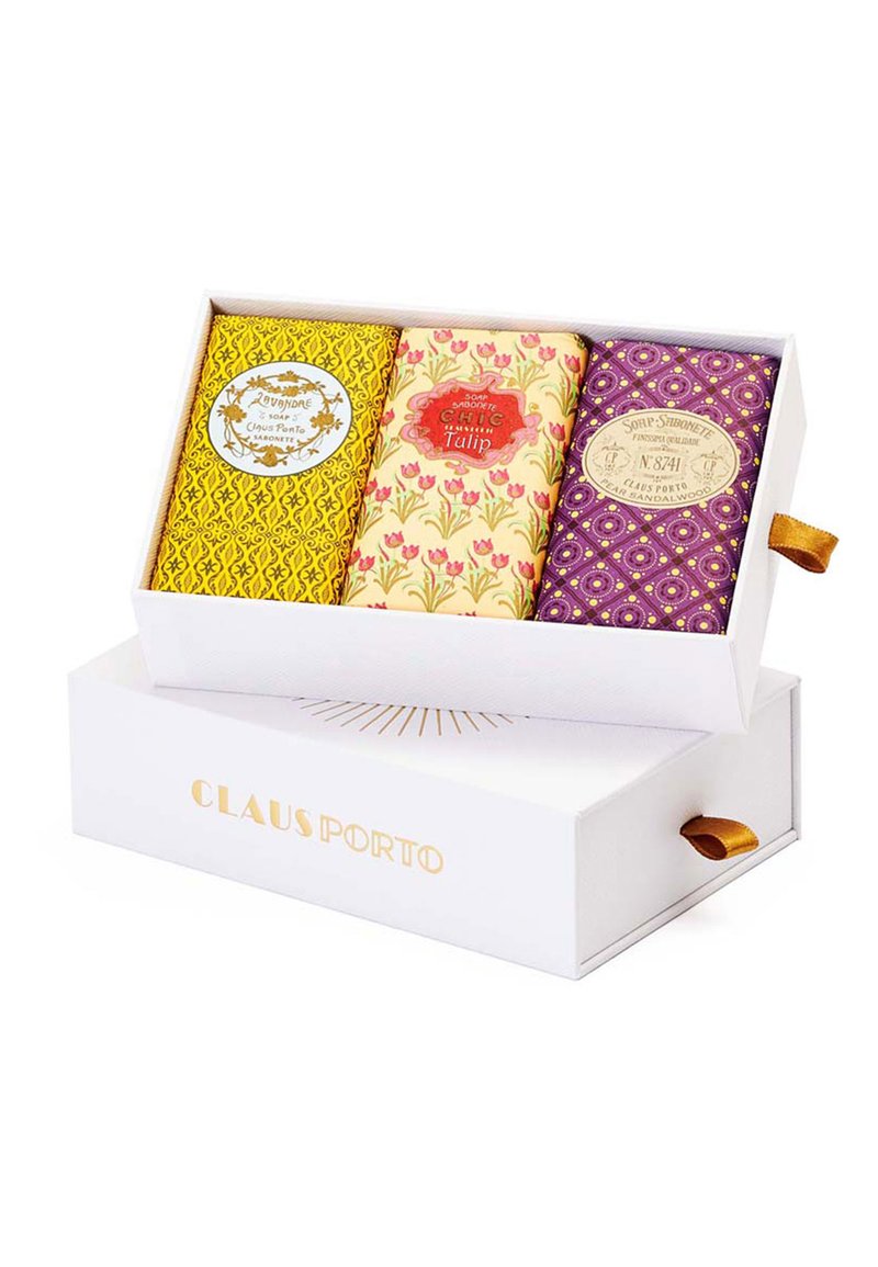 Drei dekorative Seifenstücke in einer weißen Box. Gelbe, blumige und lila gemusterte Verpackungen mit Etiketten. Die Box hat ein goldenes Logo und ein Band.