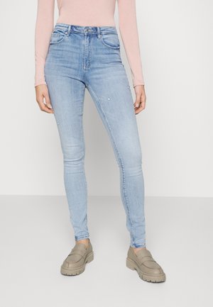 Jeans skinny azul claro com um design desbotado, dois bolsos frontais e fecho de zip. Combinados com sapatos slip-on bege.