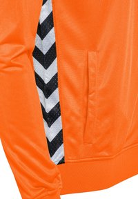 Orange sportjakke med struktureret stof, sort-hvide sidepaneler med zigzag-mønster og en diskret frontlomme.