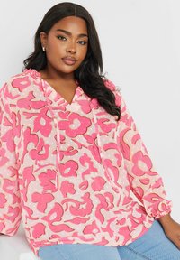 Blusa trasparente a fiori rosa con scollatura a V, cordoncini e maniche lunghe arricciate, abbinata a jeans azzurri.