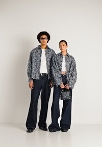 Divas modeļi, tērpti melnbaltos teksturētos žakettiem, baltās blūzēs un platveida melnās biksēs; viens tur studētu melnu rokassomu, stāvot pret vienkāršu sienu.