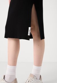 Bas des jambes portant des chaussettes blanches et des baskets beiges, présentés avec une jupe noire côtelée avec une fente latérale et une petite étiquette blanche.